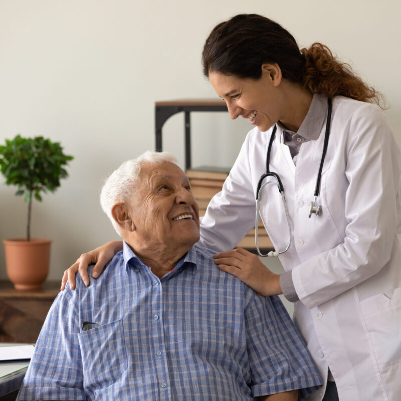 Optimistic young lady doctor embrace shoulders of laughing old man sitting on chair at clinic office
